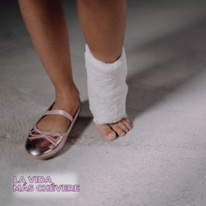A view of a pair of legs from below the knees. One wears a shinny pink ballet flat with a bow, the other is wrapped in gauze. The title of the podcast La Vida Más Chévere in bold block pink font in the corner